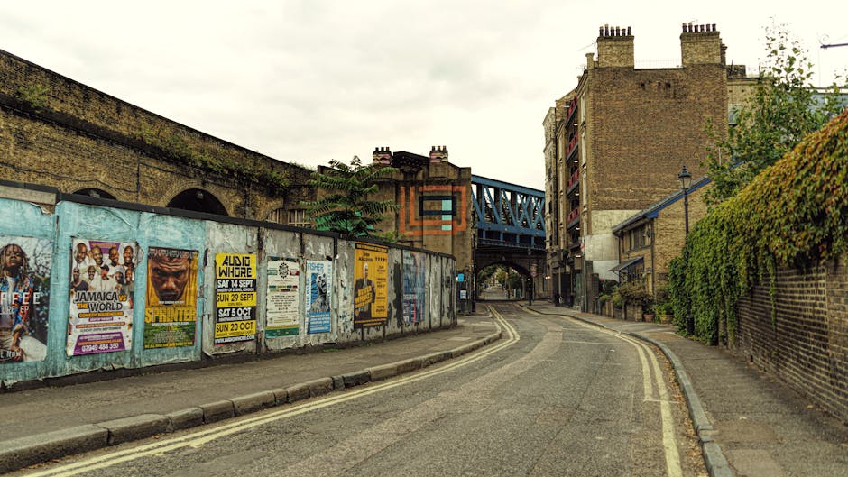 A quiet street scene near Ridley Road Market in Dalston, featuring a narrow asphalt road with double yellow lines along the curb, leading towards an underpass with a blue metal bridge. On the left side, a long row of weathered wooden and metal-panelled advertising boards display posters for local events and performances, some partially torn or faded. To the right, a brick wall covered with green foliage runs parallel to the street, with a small streetlamp mounted on a wall near the top of the right side. The scene is lit by natural daylight, with an overcast sky overhead. In the context of house removals or furniture transport, this environment illustrates a typical urban setting where a professional moving company like Man with Van Dalston could operate, loading or unloading household items at the roadside or navigating narrow pathways for a home relocation process.