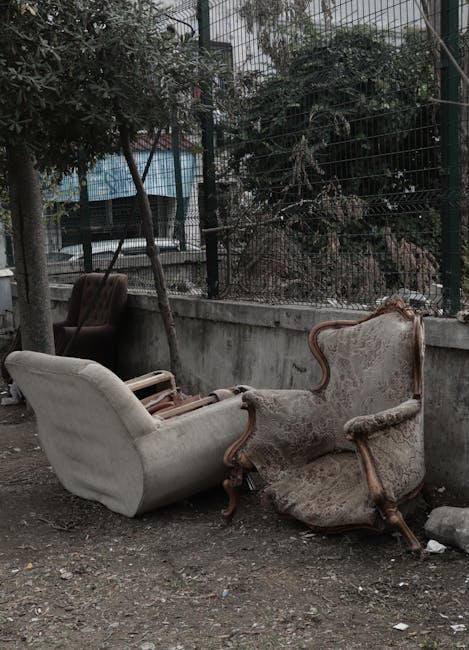 Two antique upholstered armchairs with wooden carved frames and fabric cushioning, one with a cream cover and the other with a patterned fabric, are placed outdoors on a dirt surface beside a concrete wall. The chairs appear weathered and are positioned near a metal fence with trees and urban buildings visible in the background. The scene is illuminated by natural daylight, suggesting they have been temporarily removed from a home as part of a furniture transport or packing and moving process. Man with Van Dalston specializes in removals and handling large household items, and these chairs are likely being prepared for collection or disposal during a home relocation or bulky waste removal service.