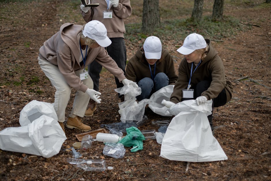 Three individuals wearing white caps, gloves, and casual outdoor clothing are outdoors on a forest floor covered with brown leaves and dirt. They are engaged in a cleanup activity, carefully collecting and placing various types of trash into large white waste bags. The trash includes plastic bottles, a green glove, and other small debris. One person is crouched down, holding a waste bag open, while the others are kneeling and reaching for items on the ground, all focused on tidying the area. An additional person stands nearby, observing or assisting with the waste collection. The scene is well-lit with natural daylight, and trees with trunks and sparse foliage are visible in the background. This outdoor waste removal activity reflects environmentally conscious efforts often associated with community cleanup events or environmental volunteering, aligned with the principles of responsible disposal and environmental preservation related to removals and waste clearance services provided by companies like Man with Van Dalston.
