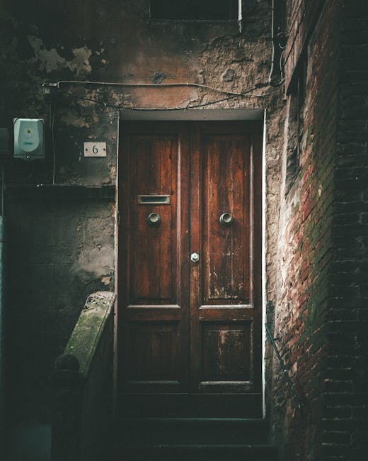 A close-up image of a narrow wooden door with a dark, weathered finish, set within an aged brick and concrete entranceway. The door features a mail slot, two round door knockers, and a small, central lock mechanism. To the left of the door, a small metal box is mounted on the concrete wall with a white number '6' plaque above it. The surrounding walls are constructed of exposed brick and rough, unpainted concrete, showing signs of wear and age, with some moss or grime present. An electrical wire runs along the top of the entrance, and a weathered stone or concrete stairway with a small ledge on the left leads up to the door. This scene is typical of urban residential entrances in a city setting, often encountered during home relocations or furniture transport processes, where narrow entrances require careful maneuvering by professional removals services like Man with Van Dalston.