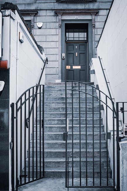 A set of concrete stairs leading up to a dark green front door with a windowpane at the top, situated within a grey stone building facade. Metal railings run along both sides of the stairs, and a black metal gate with a curved top secures the entrance. To the left, a white wall with what appears to be an outdoor light fixture is visible, while to the right, part of a white boundary wall extends alongside the steps. The entrance area is well-lit, and the building has a classic architectural style. This scene depicts the exterior of a residential property prepared for house removal or relocation, with the stairs and entrance ready for the loading process. Man with Van Dalston specializes in such house moves, including navigating narrow or challenging entrances like this one, ensuring the safe transport of furniture and packing materials during home relocation.