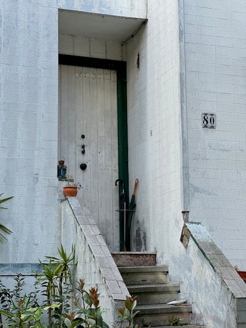 A close-up image of a narrow wooden door with a dark, weathered finish, set within an aged brick and concrete entranceway. The door features a mail slot, two round door knockers, and a small, central lock mechanism. To the left of the door, a small metal box is mounted on the concrete wall with a white number '6' plaque above it. The surrounding walls are constructed of exposed brick and rough, unpainted concrete, showing signs of wear and age, with some moss or grime present. An electrical wire runs along the top of the entrance, and a weathered stone or concrete stairway with a small ledge on the left leads up to the door. This scene is typical of urban residential entrances in a city setting, often encountered during home relocations or furniture transport processes, where narrow entrances require careful maneuvering by professional removals services like Man with Van Dalston.