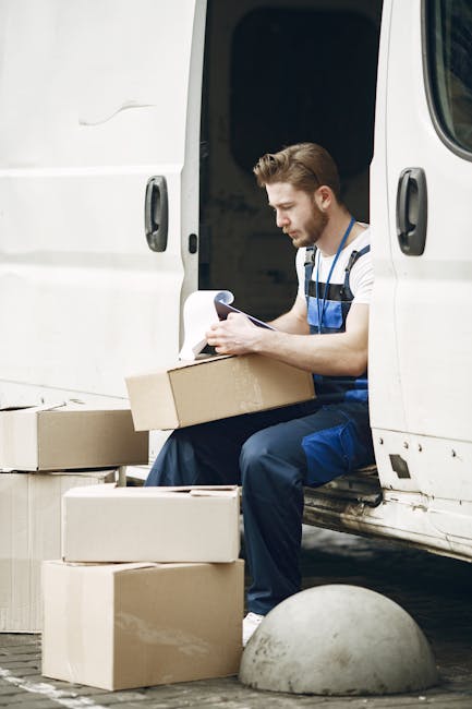 A man wearing a striped t-shirt and blue work overalls is seated on the open door of a white moving van, engaged in packing or organizing cardboard boxes for a home relocation. Several packed cardboar