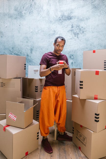 A man wearing a striped t-shirt and blue work overalls is seated on the open door of a white moving van, engaged in packing or organizing cardboard boxes for a home relocation. Several packed cardboar