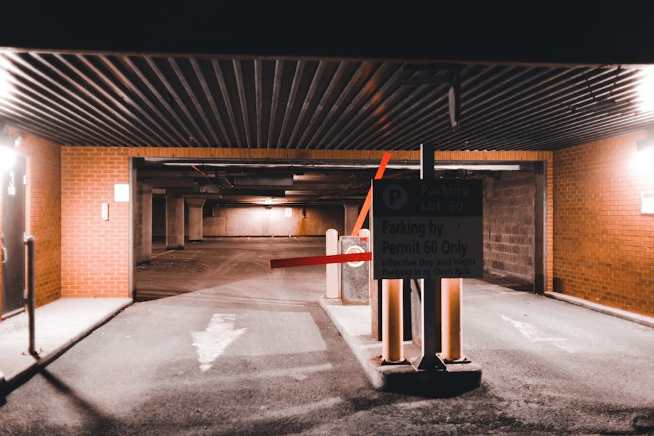 Inside a multi-story parking garage with brick walls and a low, dark ceiling with wooden slats, a parking barrier gate is seen blocking the entrance. The barrier arm is red and positioned horizontally. A parking sign attached to the barrier indicates parking lot 60 and permission restrictions, likely relevant to house removals or relocation logistics in Dalston, as referenced by the page title. The area is illuminated by artificial lighting, casting a warm glow on the concrete floor marked with a white arrow guiding vehicles towards the exit or entrance. To the left, the edge of a doorway or wall is visible, possibly leading to an indoor storage or loading area. The scene reflects a typical environment for loading furniture, packed boxes, and removal items during an internal or external home relocation. Man with Van Dalston's team may use this space during the loading process for furniture transport, packing, or moving services, with vehicles positioned nearby for swift transfer of household belongings.
