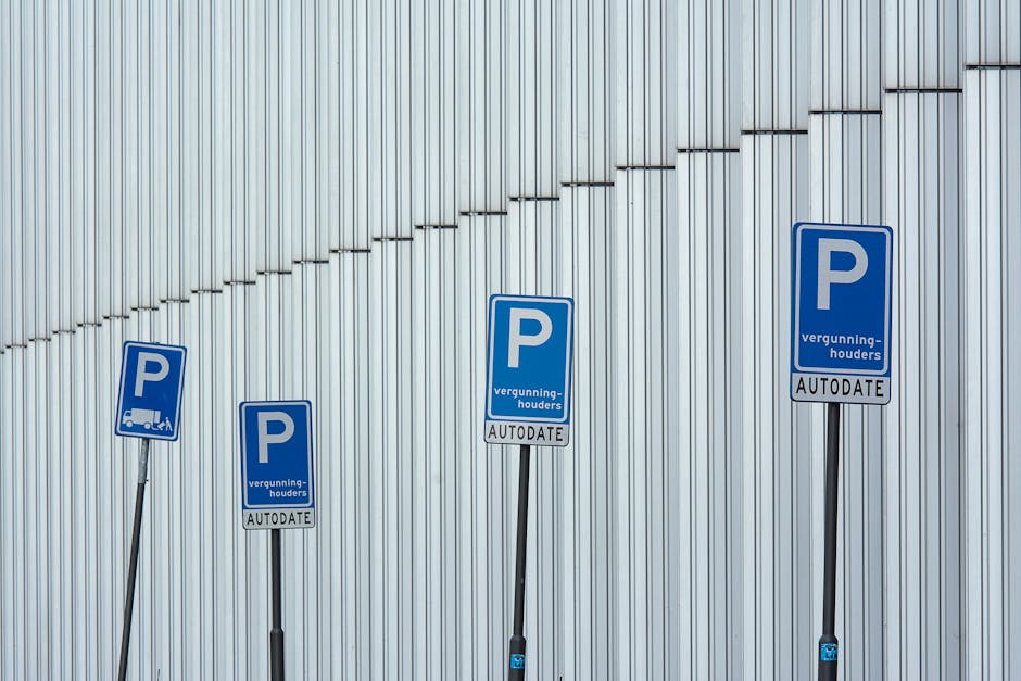 Four blue parking permit signs mounted on black metal poles are positioned in front of a vertical metallic wall with a series of small, evenly spaced protrusions forming a pattern. The signs display a white capital 'P' and the words 'vergunning-houders' underneath, with the word 'AUTODATE' in a smaller font at the bottom. The signs are arranged in a slightly diagonal line, with the leftmost sign leaning slightly to the left and the rightmost sign positioned upright. The environment appears to be an outdoor area, possibly near a building used for house removals or moving logistics, with natural daylight illuminating the scene. The signs are likely related to parking permits for vehicles involved in local relocations, supporting the context of house removal services offered by Man with Van Dalston, as indicated on the page about Hackney Council parking permits for Dalston movers.