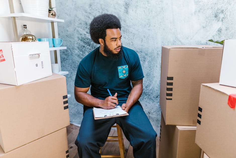 A man with curly black hair and a beard is sitting on a small wooden stool inside a room with a textured, light blue wall. He is wearing a dark blue T-shirt with a patterned pocket on the left side and dark trousers. The man is holding a blue pen in his right hand and writing on a clipboard resting on his lap, appearing to be assessing or planning a move. Surrounding him are several cardboard boxes, some sealed with black tape and others open or partially packed, positioned on the floor and stacked against the wall. Behind him, a white shelving unit holds various decorative objects, including a glass bottle, vases, and cups. The room is well-lit, likely from natural light, indicating a typical home environment. This scene exemplifies the process of home relocation or furniture transport preparation, with [COMPANY_NAME] providing professional removals services, as suggested by the context.