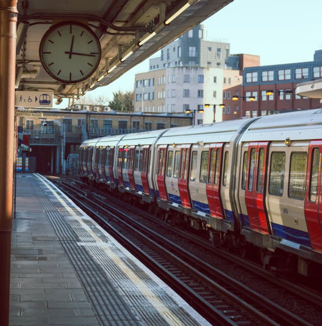 The image depicts an empty London underground train station platform at night, illuminated by overhead lighting. The platform has a row of black and yellow safety bollards along the edge, and tactile paving near the platform edge for visually impaired passengers. On the right side, there are brick walls with several windows and a round London Underground sign affixed to one of the columns supporting the roof. The station’s canopy is supported by decorative columns with yellow accents, and there are benches and a few posters visible along the platform. The tracks run parallel to the platform into a dark tunnel at the far end. The scene is clean and well-maintained, suggesting a setting where house removals or furniture transport could be coordinated, such as loading or unloading items for a home relocation, with the station as part of transport logistics managed by [COMPANY_NAME].