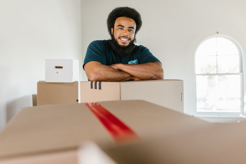 A smiling man with dark, curly hair and a beard stands inside a well-lit room, leaning on a collection of packed cardboard moving boxes of various sizes, some sealed with red tape. The boxes are arranged on the floor and ledge, indicating a home relocation process. Behind him, there is a white wall, and a large, arched window allowing natural light to fill the space. The scene captures the preparation stage of furniture transport and packing for a house move, with the man from Man with Van Dalston overseeing the packing and loading process, which involves careful handling of boxes in the indoor environment prior to transport to a new property.