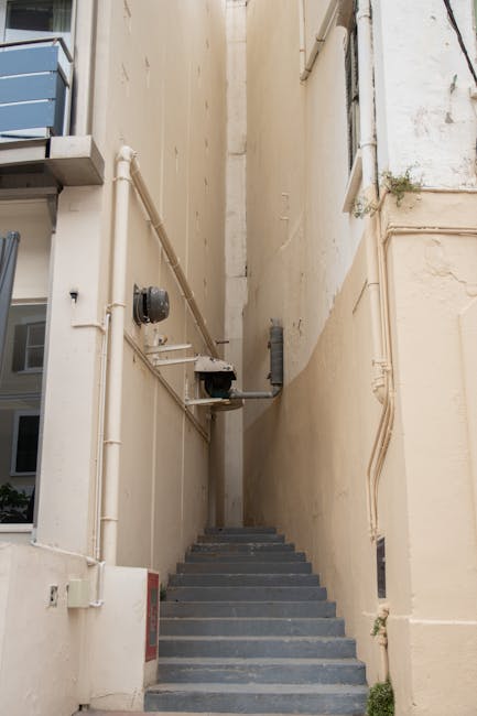 A narrow exterior staircase with dark grey steps leading upward between two beige-faced buildings, surrounded by utility pipes and cables attached to the wall on the left side. The staircase appears to be part of a residential property, possibly an alleyway or side access to a home. The lighting is natural, suggesting daytime. In the background, the upper portions of the neighboring building are partially visible. This scene depicts the type of access involved in house removals or furniture transport during a home relocation, which Man with Van Dalston can assist with as part of their removals services.