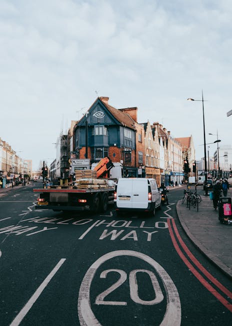 A flatbed truck with an open cargo area is parked on a busy street in Dalston during a house removal process, operated by Man with Van Dalston. The truck is loaded with wooden pallets and cardboard boxes, some wrapped in plastic and fabric blankets to protect fragile items. The vehicle is positioned partially in the middle of the road, behind a white van. The scene shows a row of multi-story buildings with a mix of brick and painted facades, typical of Stoke Newington Road, with pedestrians, parked bicycles, and street lamps along the pavement. The street markings indicate a 20 mph speed limit and designated lanes for vehicles and bicycles. The overall environment suggests a relocation, with furniture and packing materials prepared for transfer, while the loading process appears to be ongoing, illustrating the careful handling typical of professional moving services such as those provided by Man with Van Dalston in the context of tight access in Dalston.